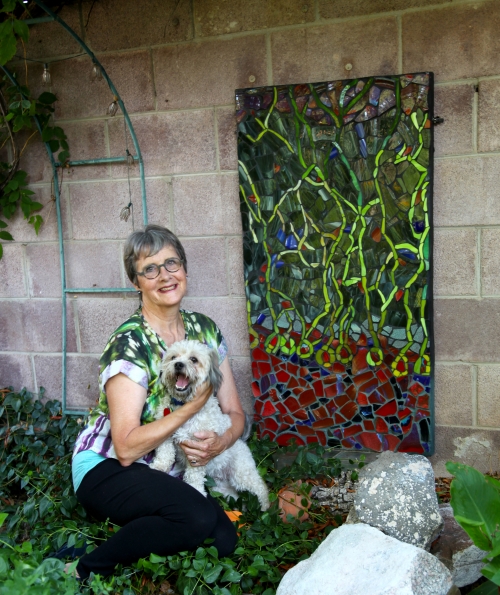 A woman holding a small dog, kneeling in a garden next to a colorful mosaic.